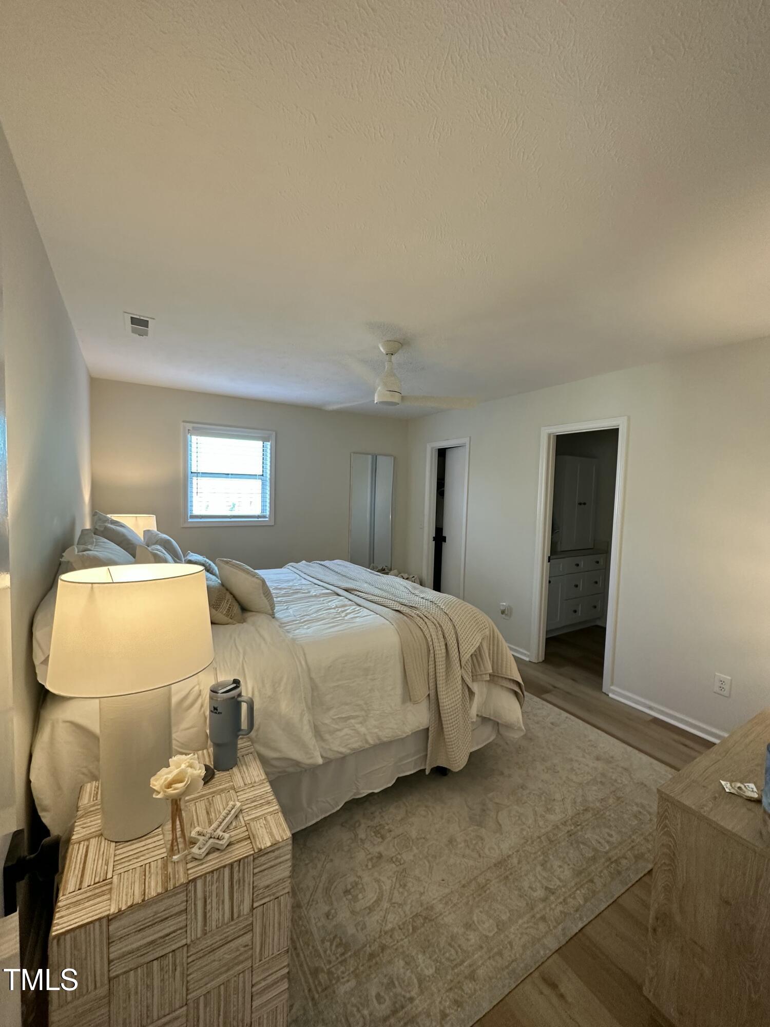 Furnished bedroom with a large neutral-toned bed, bedside lamp, ceiling fan, and patterned rug.