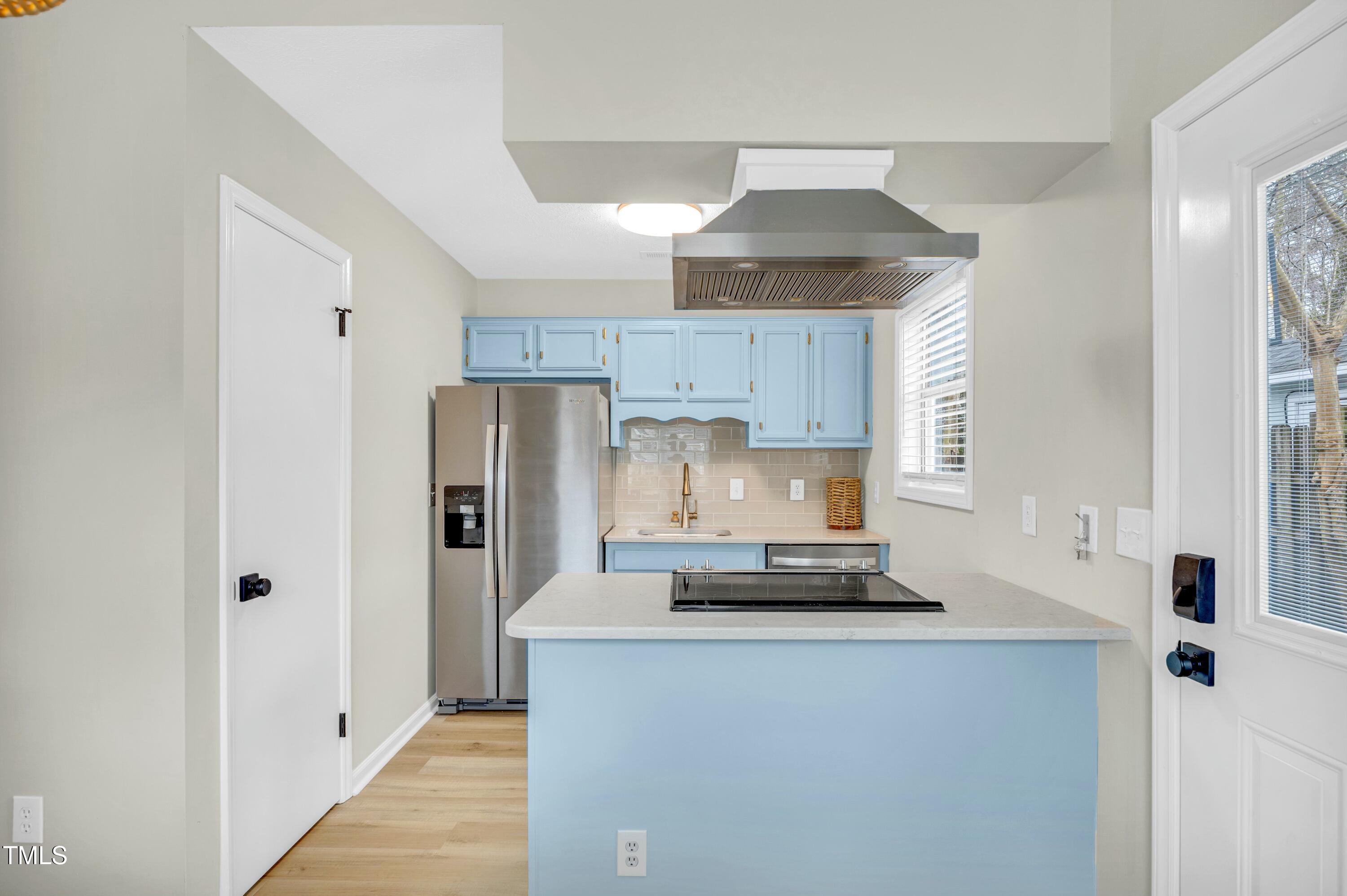 Kitchen with light blue cabinets and island, stainless appliances, and a cooktop beneath a range hood.