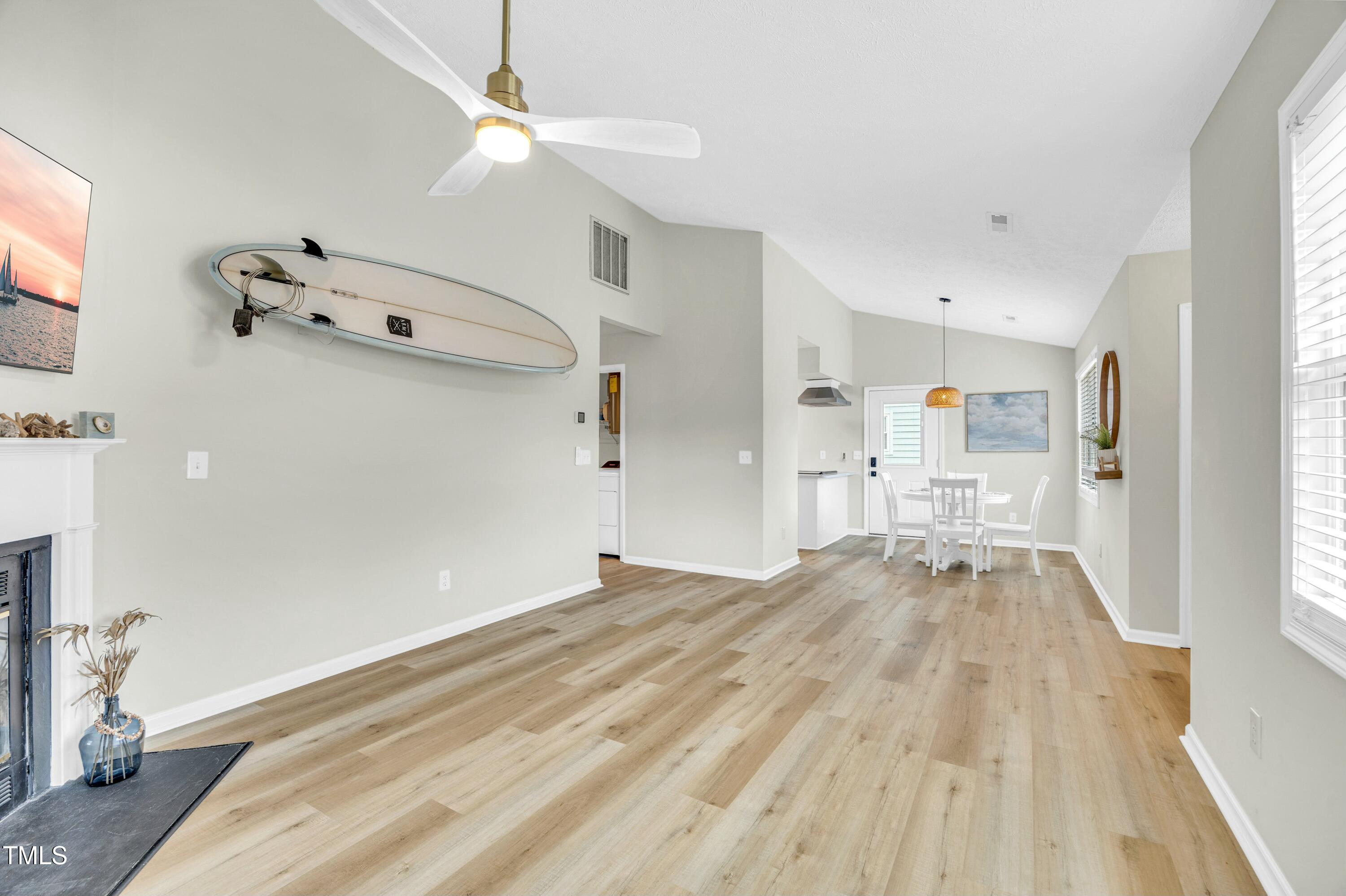 Open-plan living and dining room with a vaulted ceiling, mounted surfboard, fireplace, and light wood floors.