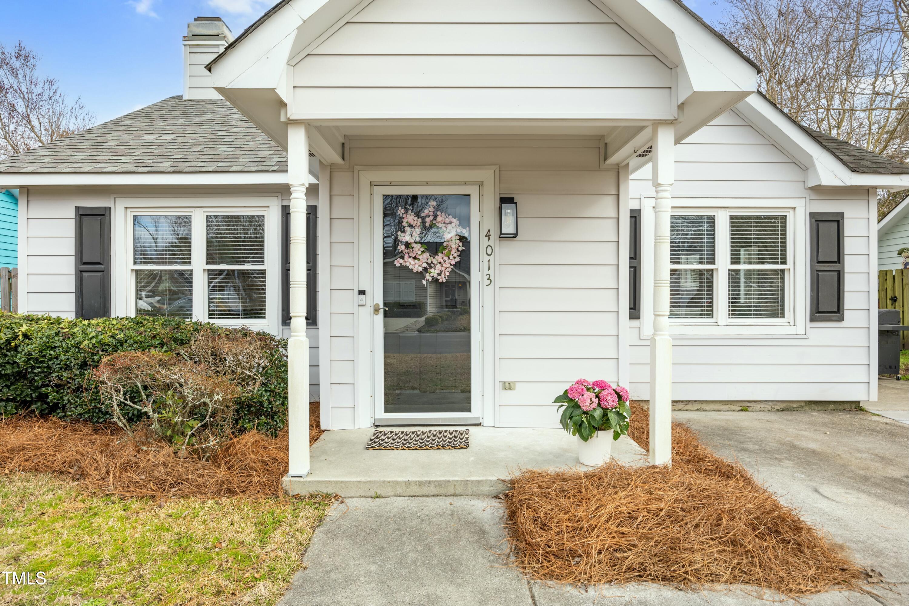 White single-story home with a covered porch, floral wreath on the glass door, and pink flowers by the entry.