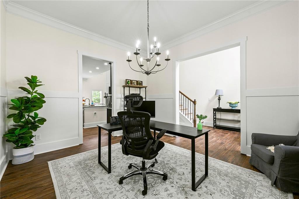 Bright home office with a black desk, ergonomic chair, chandelier, and doorways to the kitchen and stairs.