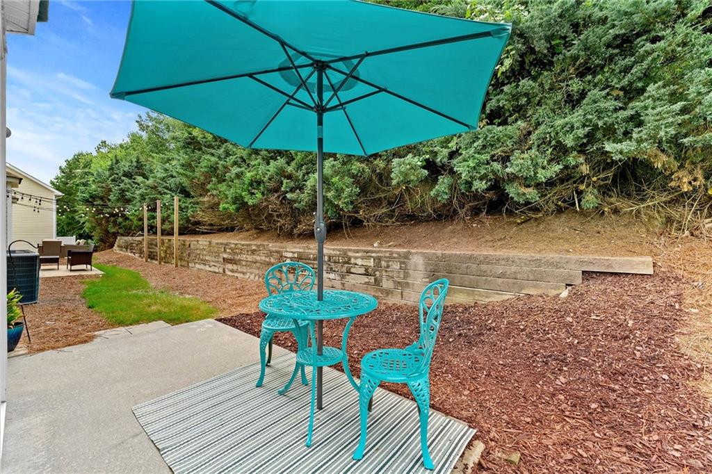 Backyard patio with turquoise umbrella and metal seating beside a mulched yard and retaining wall.