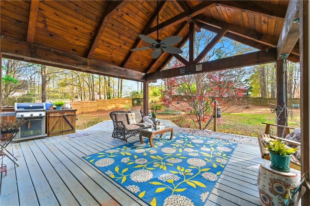 Covered backyard deck with exposed wooden beams, outdoor seating, and a blue floral rug.