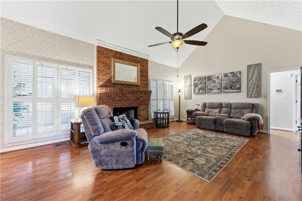 Living room with a brick fireplace, vaulted ceiling, plantation shutters, and hardwood floors.
