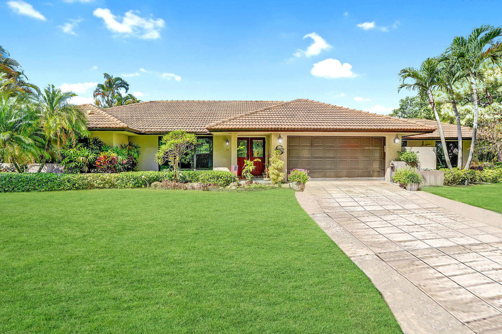 Single-story home with a tile roof, red front door, double garage, tropical landscaping, and broad green lawn.