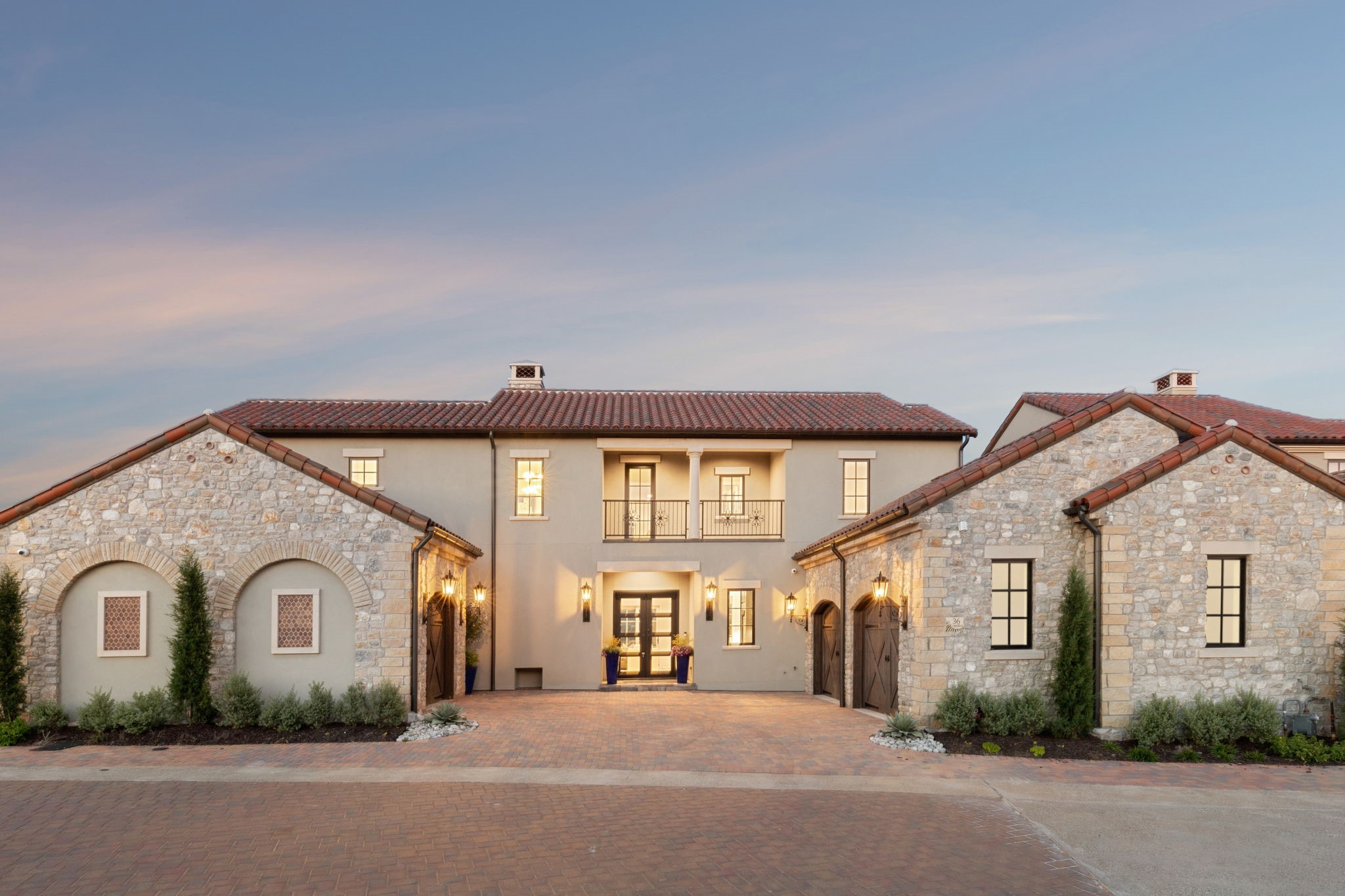 Two-story stone and stucco home with a central balcony and broad brick courtyard at dusk.