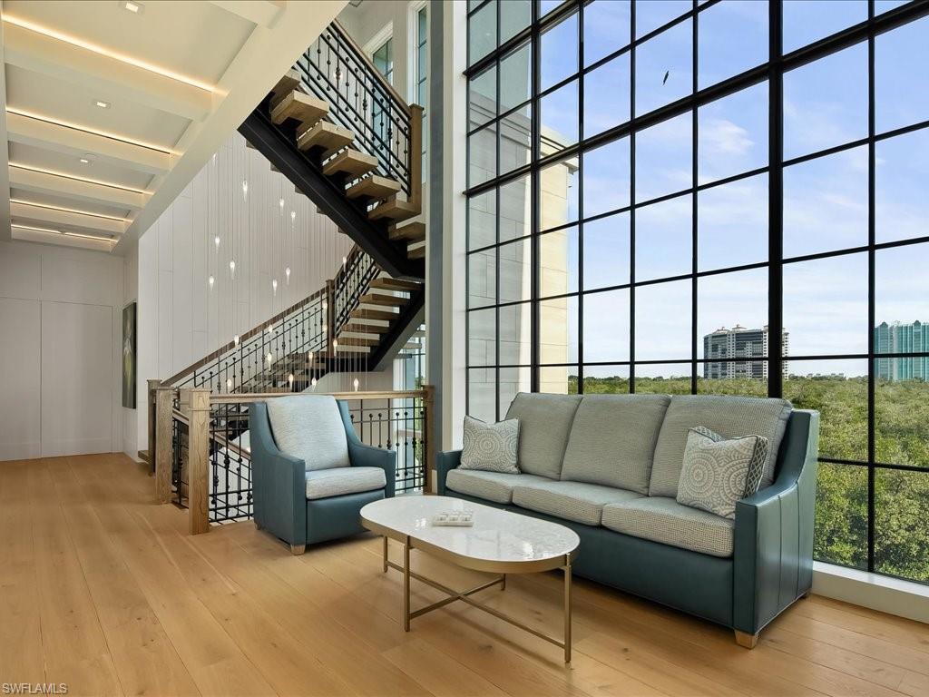 Furnished living area with a dramatic staircase and floor-to-ceiling windows overlooking greenery.
