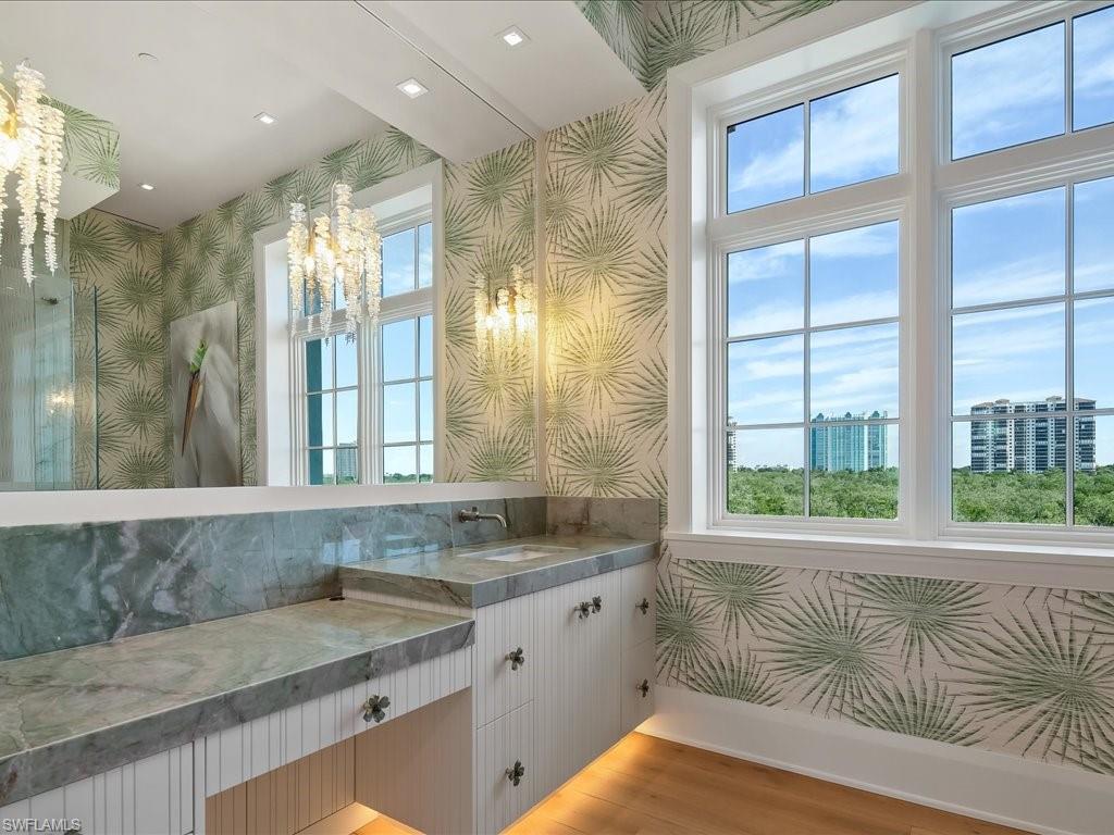 Bathroom vanity with marble counters, palm-leaf wallpaper, and tall windows overlooking greenery and high-rises.