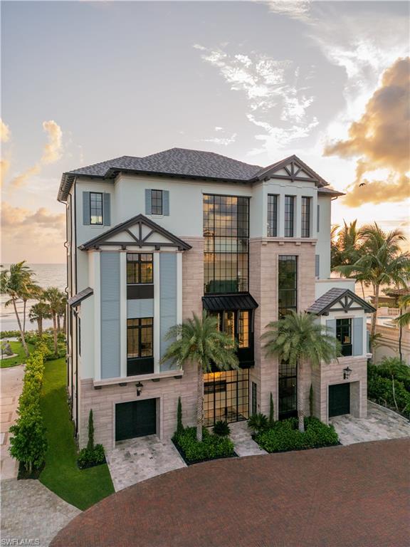 Modern coastal home with tall central windows, palm trees, and water visible beside it.