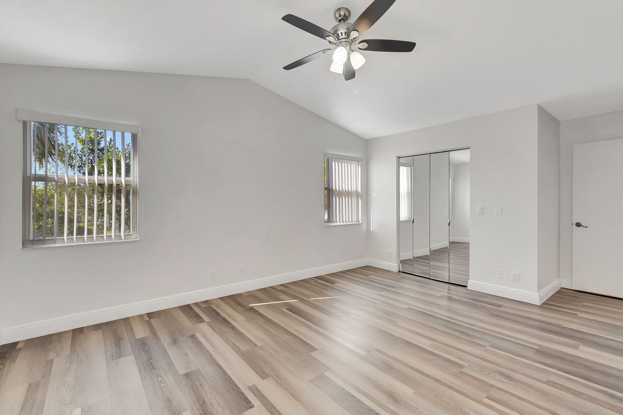 Vacant bedroom with vaulted ceiling, ceiling fan, mirrored closet doors, and plank flooring.