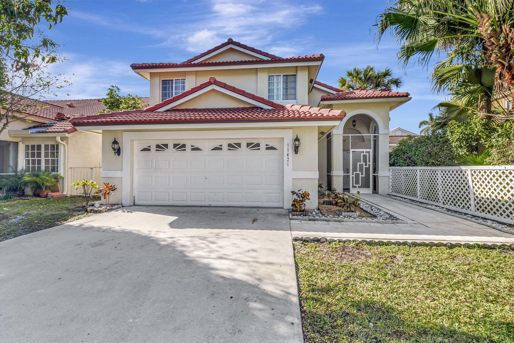 Two-story cream stucco house with red tile roofs, an arched entryway, and tropical landscaping.