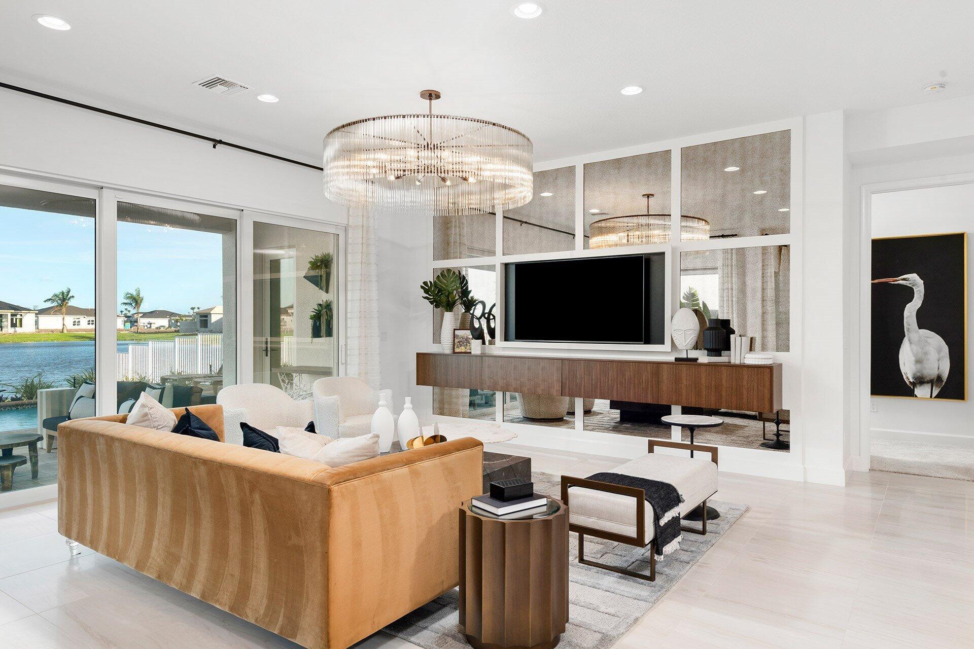Living room with a tan sofa, mirrored TV wall, and sliding doors overlooking a pool and water.