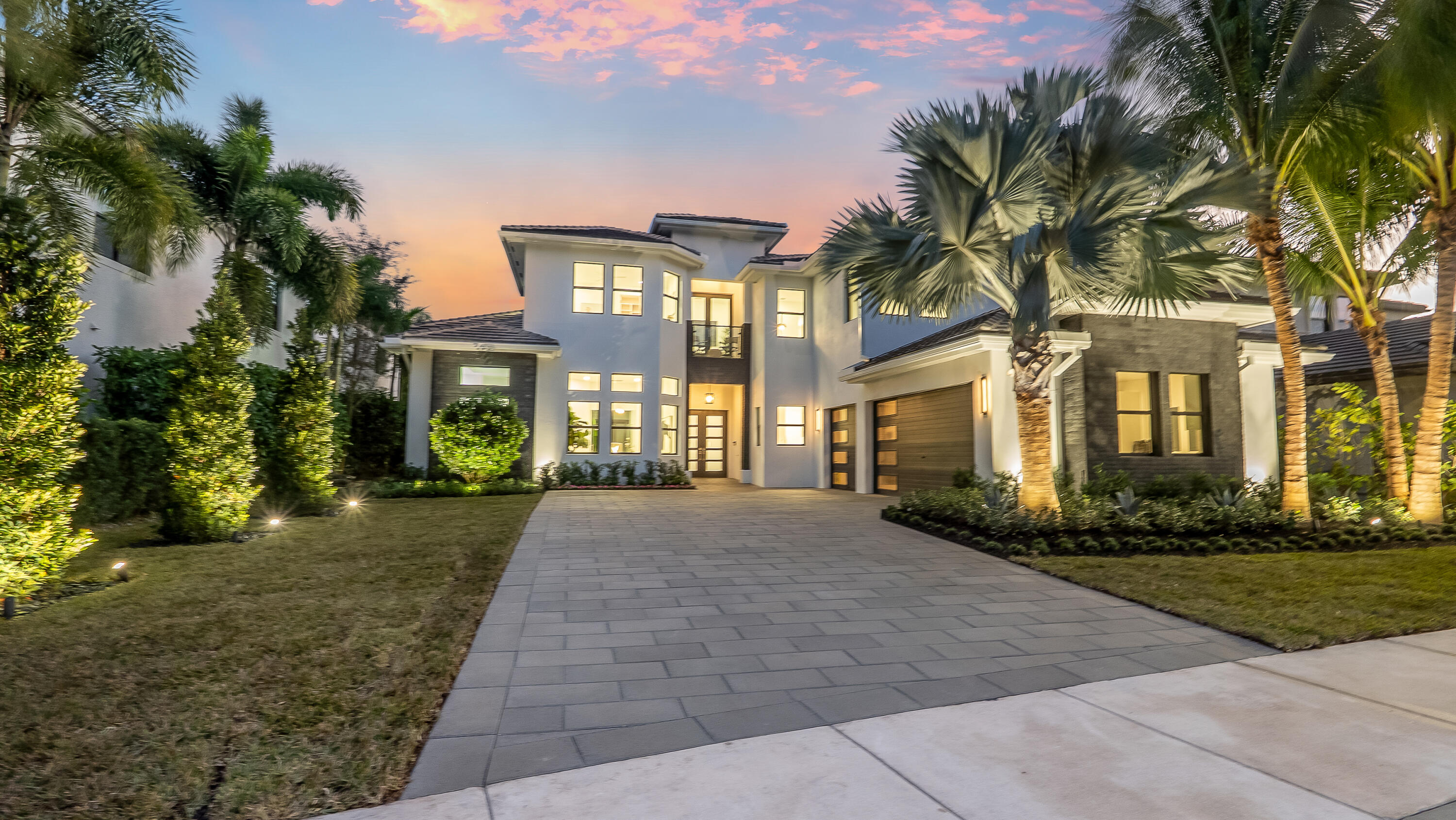 Modern two-story home with illuminated windows, a broad paved driveway, and palm trees at dusk.