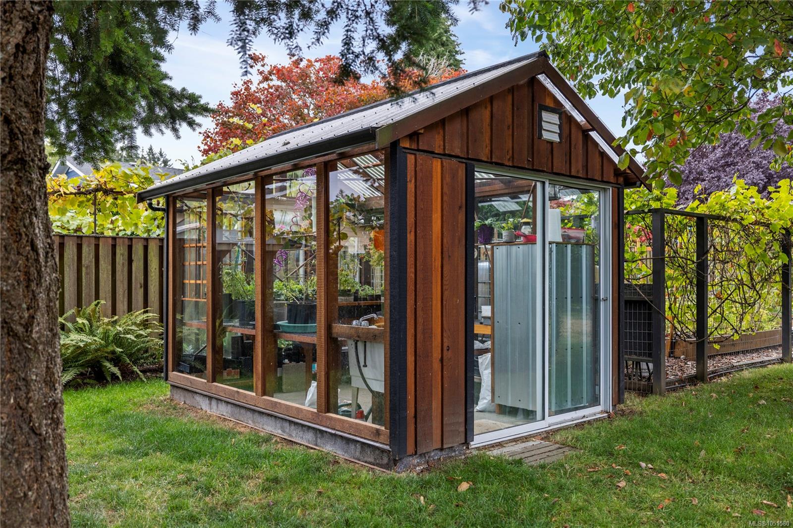 Wood-framed glass greenhouse surrounded by lawn, trees, and garden plants.