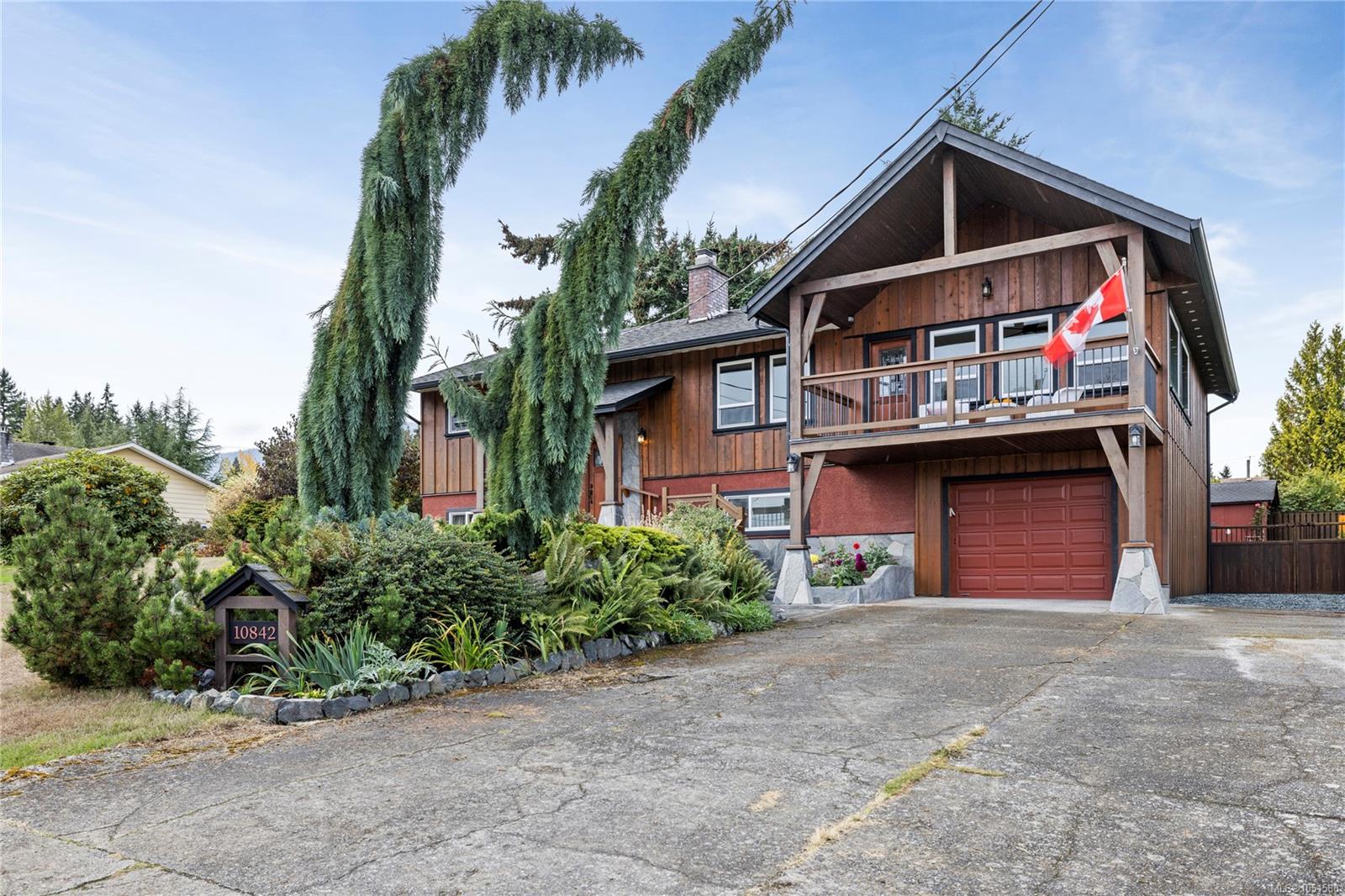 Wood-sided home with a raised balcony, Canadian flag, red garage door, and landscaped front garden.