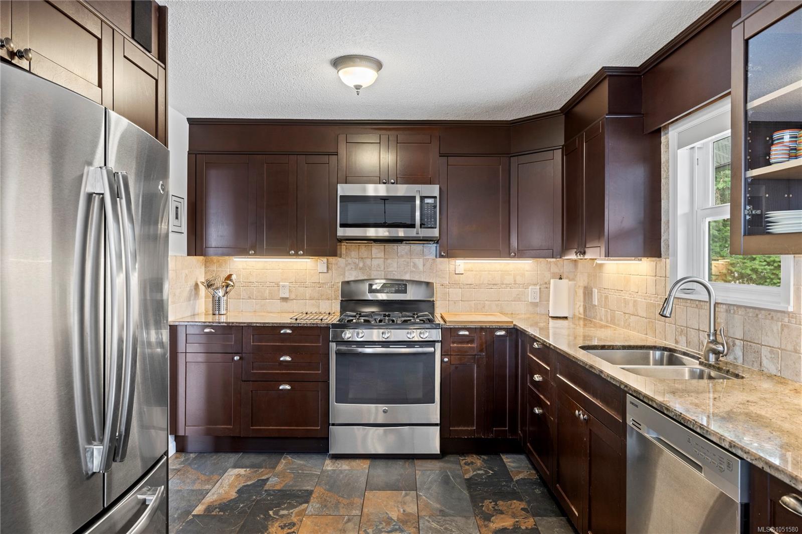 Kitchen with dark wood cabinets, stainless steel appliances, stone tile backsplash, and a double-basin sink.