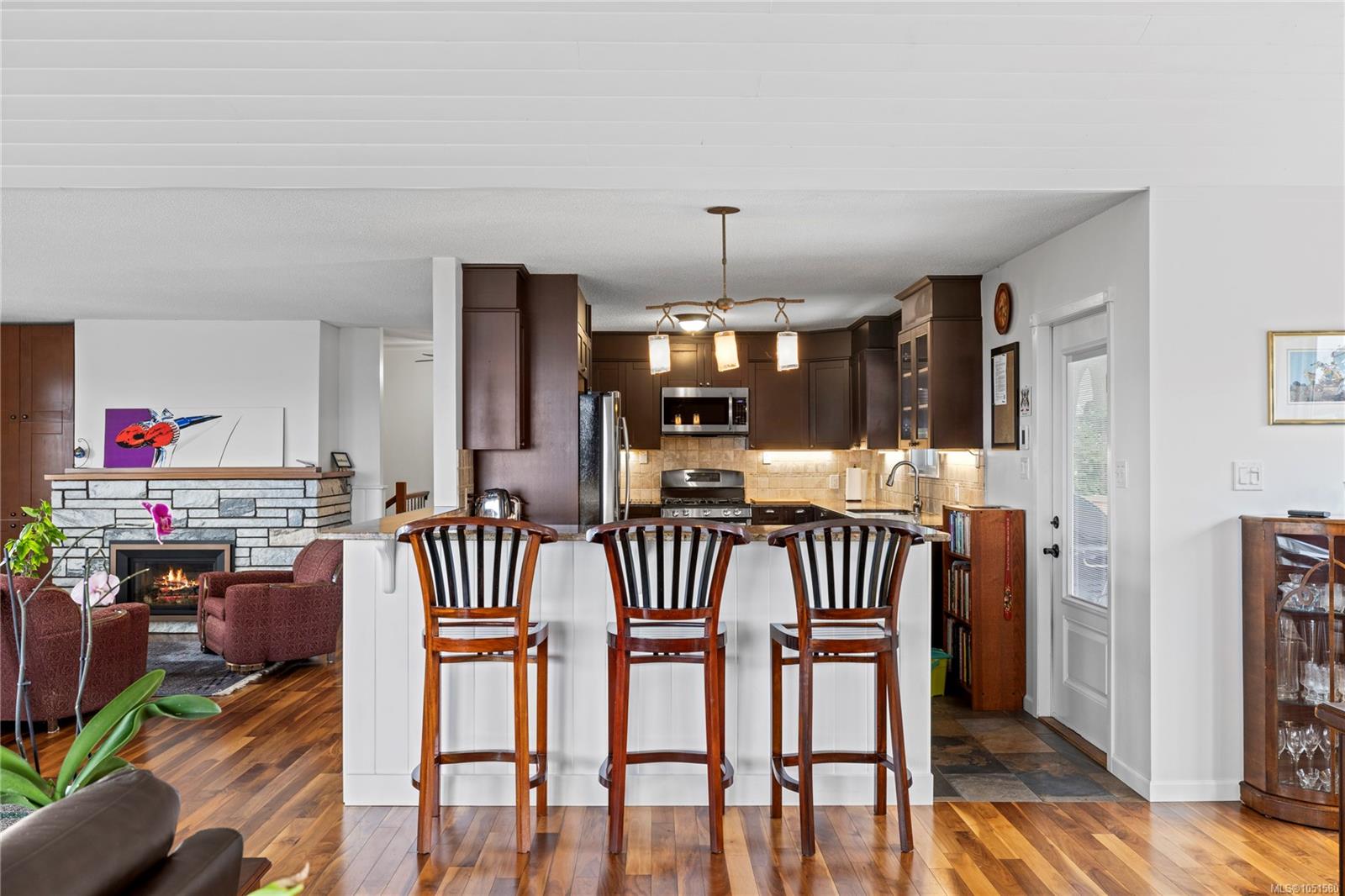 Open kitchen and living area with a white breakfast bar, dark cabinetry, and stone fireplace.