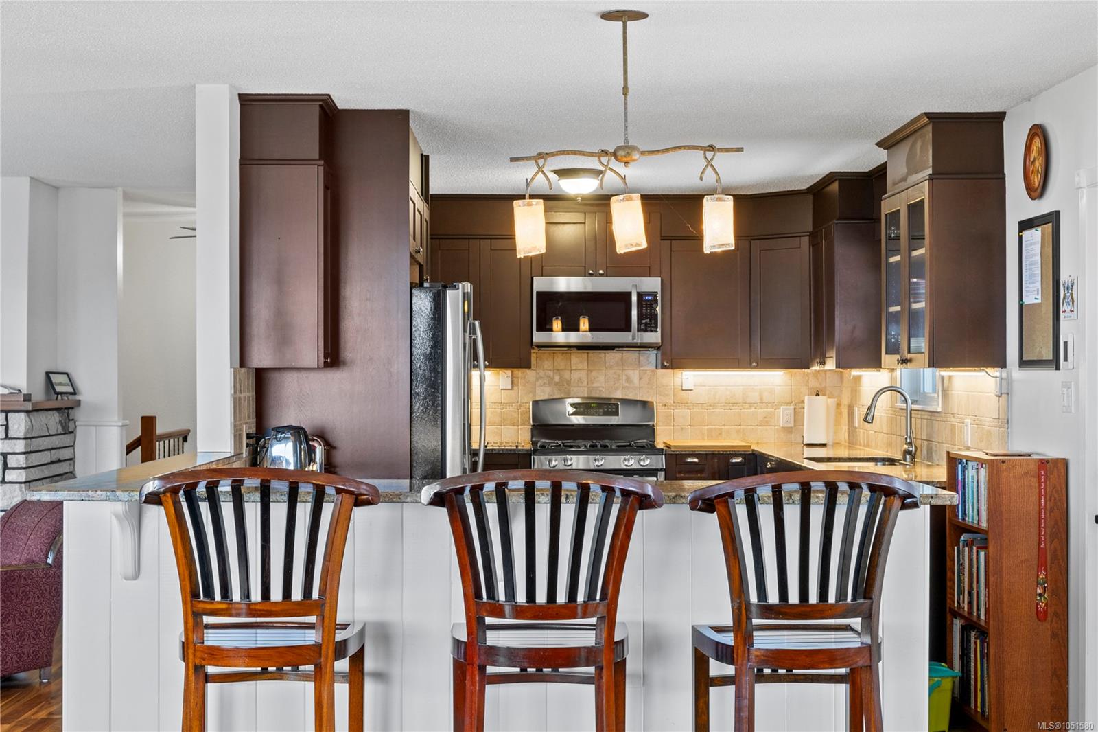 Kitchen with dark wood cabinets, tiled backsplash, stainless appliances, and a breakfast bar with three chairs.