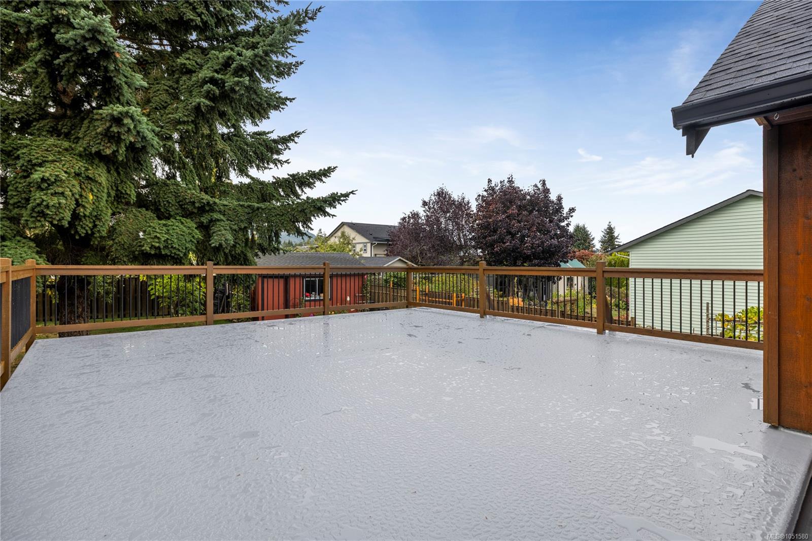 Large flat deck with brown-and-black railing overlooking trees and neighboring homes.