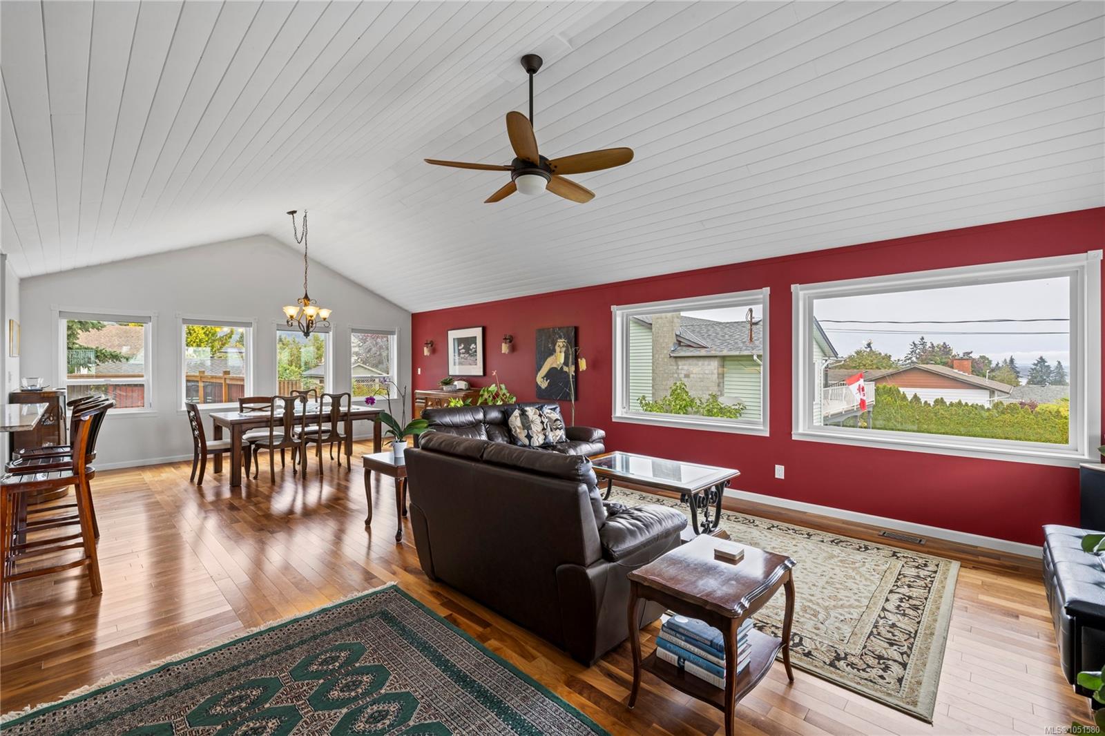 Living and dining room with vaulted ceiling, red accent wall, hardwood floors, and large windows.
