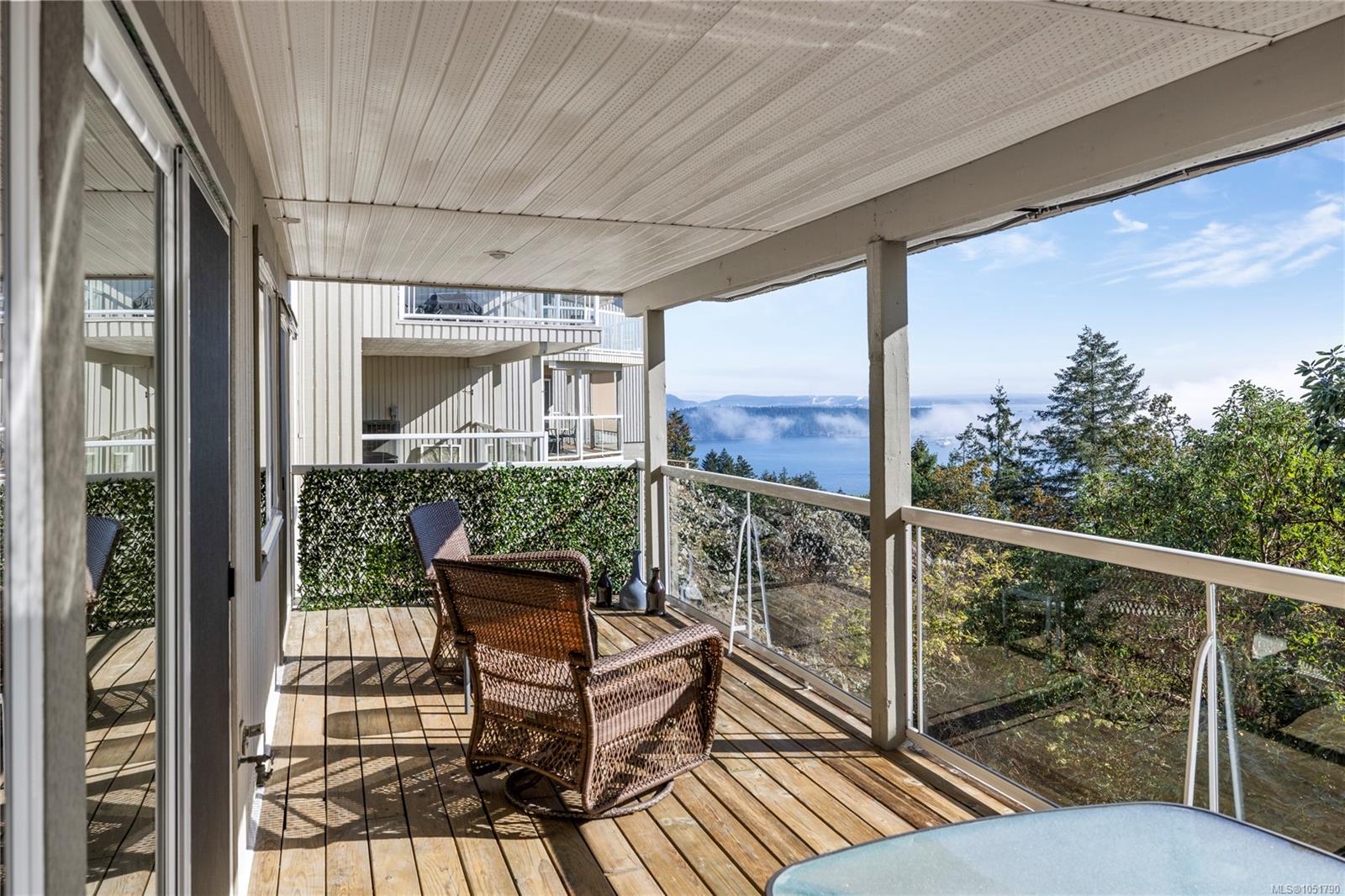 Covered deck with wicker chairs and glass railings overlooking an inlet and wooded hills.