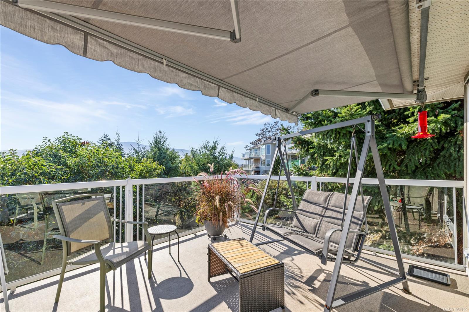 Furnished deck with a retractable awning, patio swing, glass railing, and leafy surroundings.