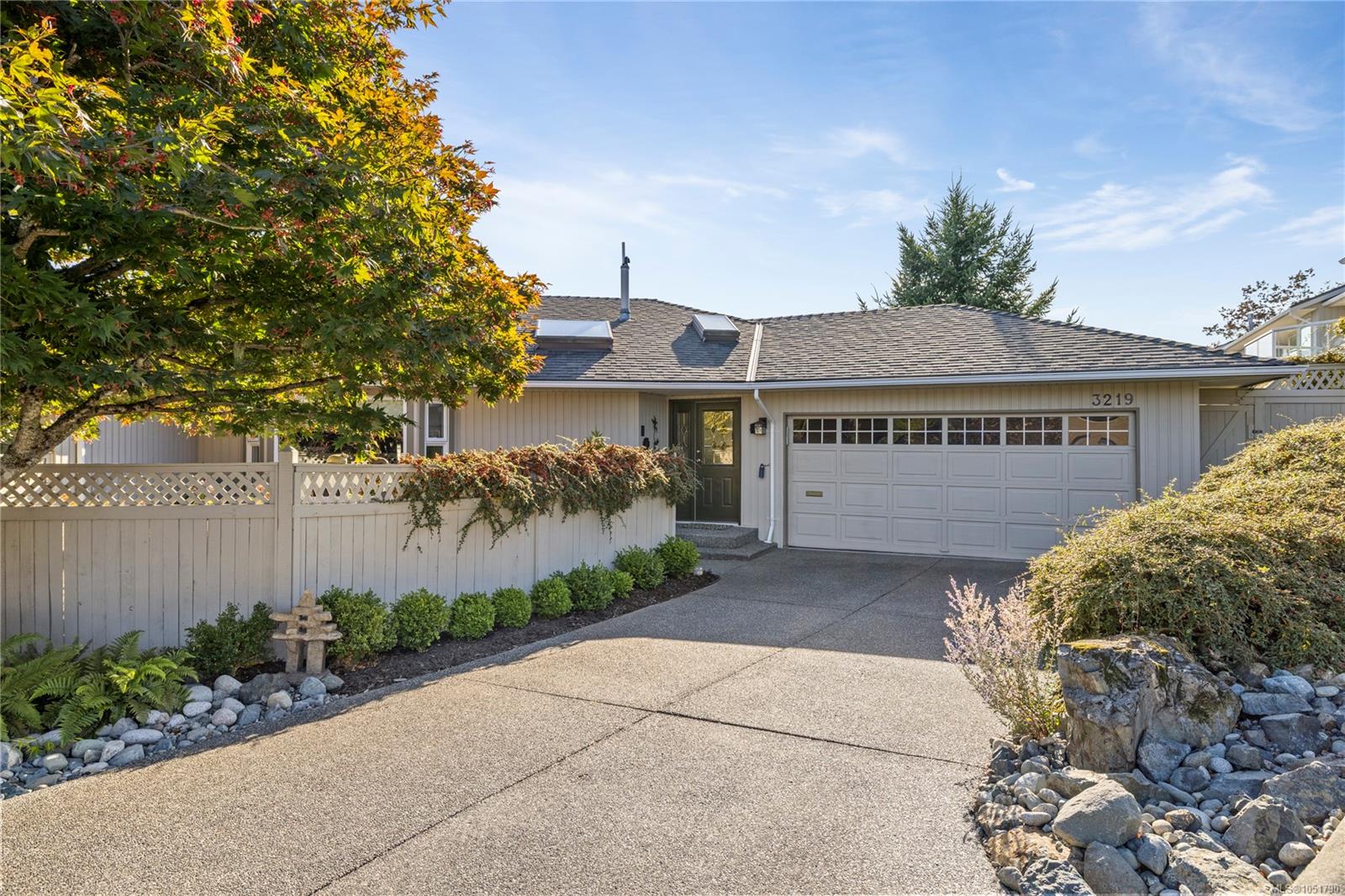 Single-story home with a double garage, broad driveway, white fence, and autumn-colored tree.