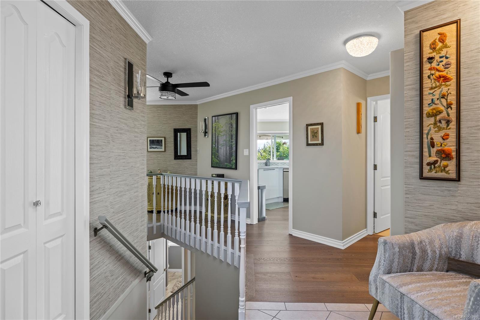 Hallway landing overlooking a staircase, with framed artwork and a bright kitchen beyond.