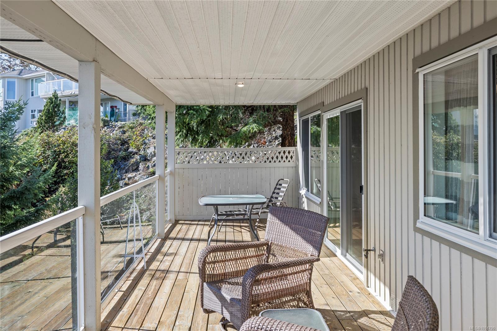 Covered wooden deck with wicker seating, glass railings, and views of a landscaped slope.