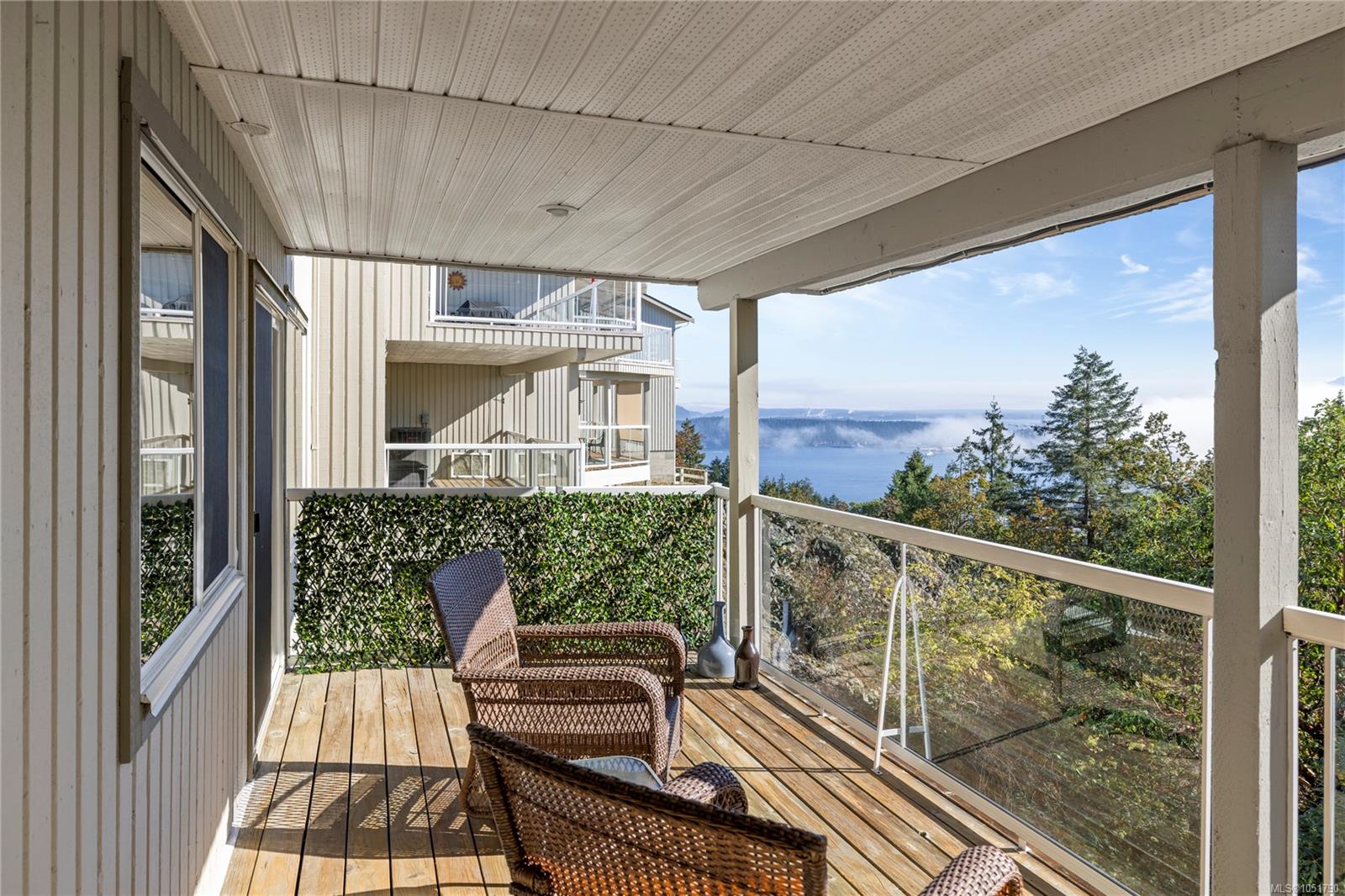 Covered balcony with wicker chairs and glass railing overlooking water and distant hills.