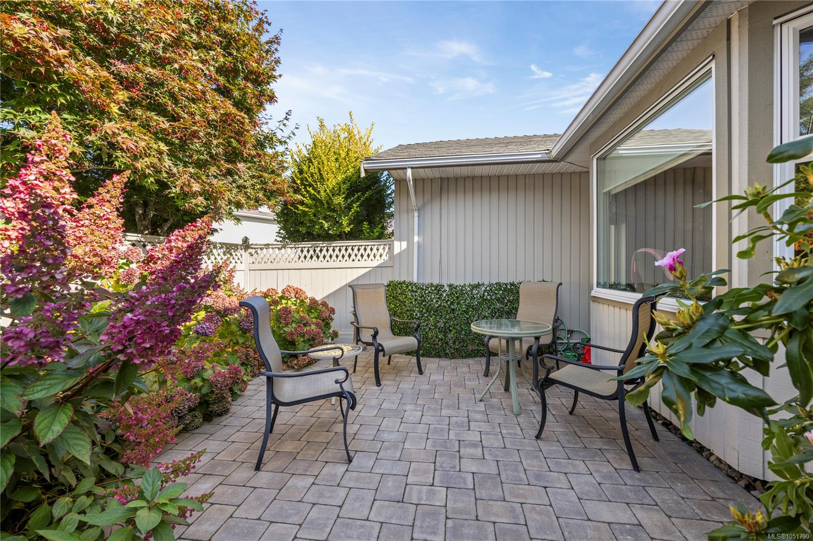 Paved patio with metal seating, glass tables, and flowering shrubs beside the house.