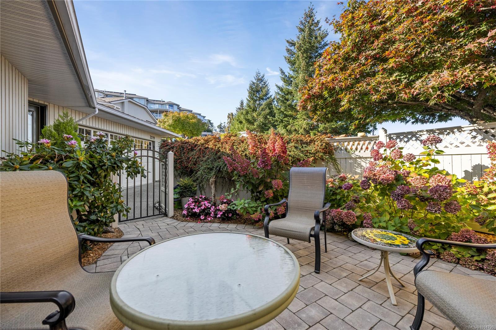 Paved patio with outdoor chairs and tables, flowering shrubs, and a white lattice fence beside the house.