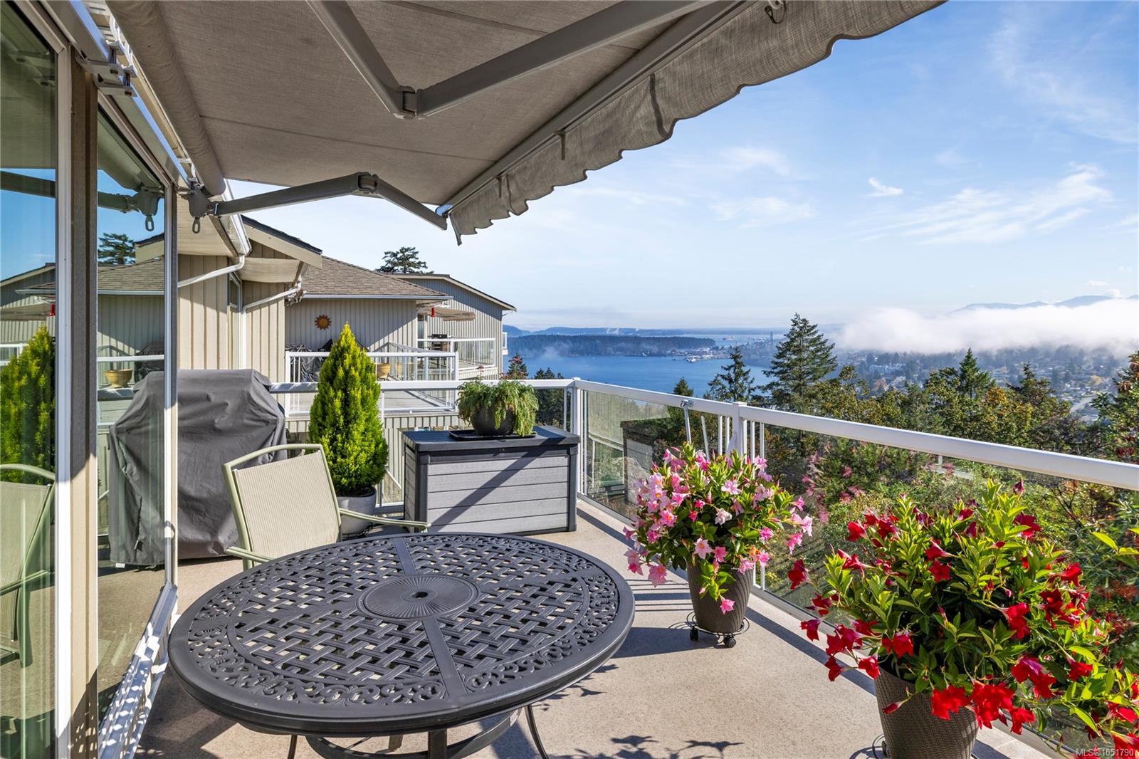 Furnished deck with flowering planters and a glass railing overlooking the water and coastal hills.
