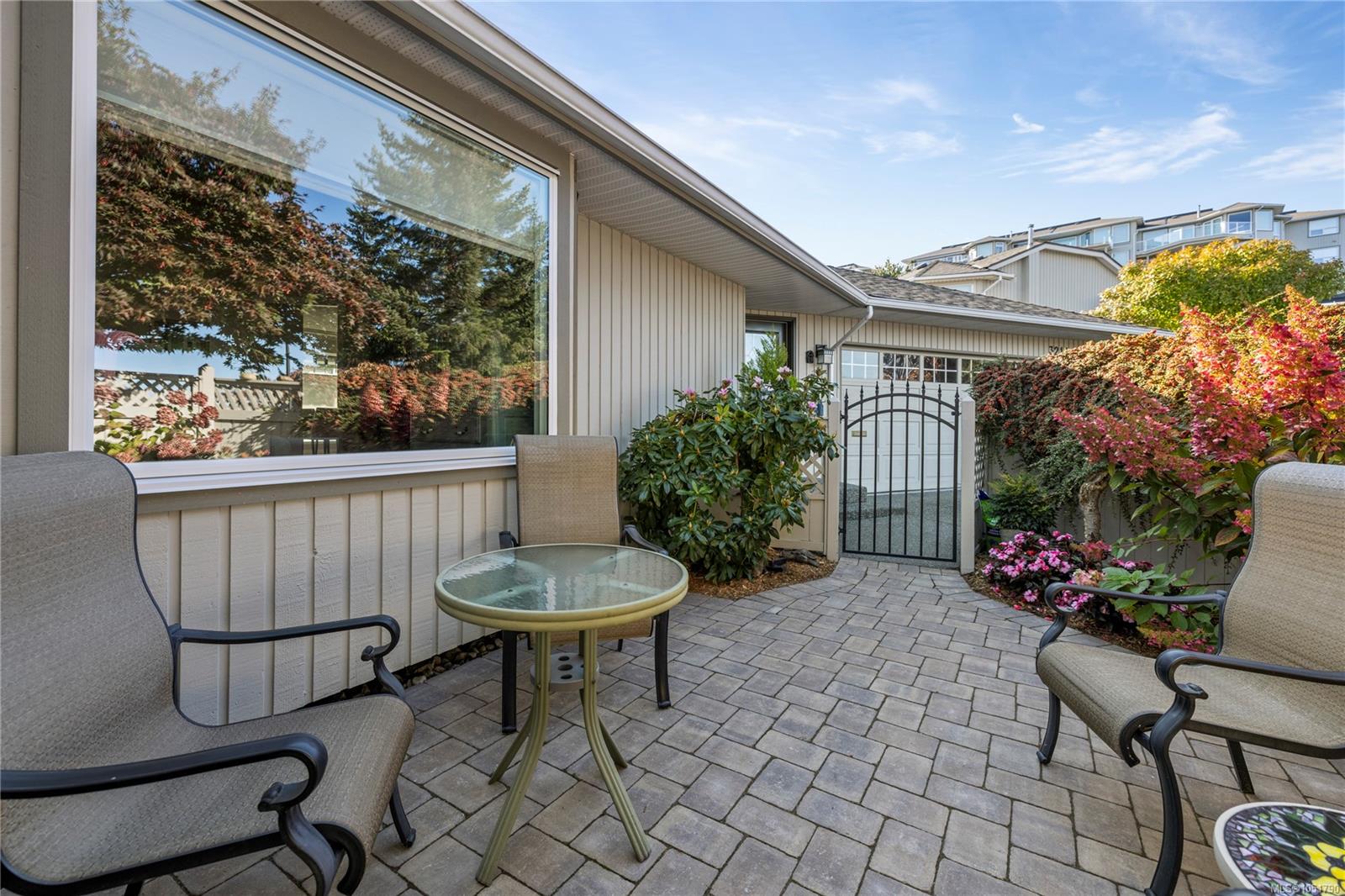 Paved patio with cushioned chairs, a glass-top table, flowering shrubs, and a black gate beside the home.