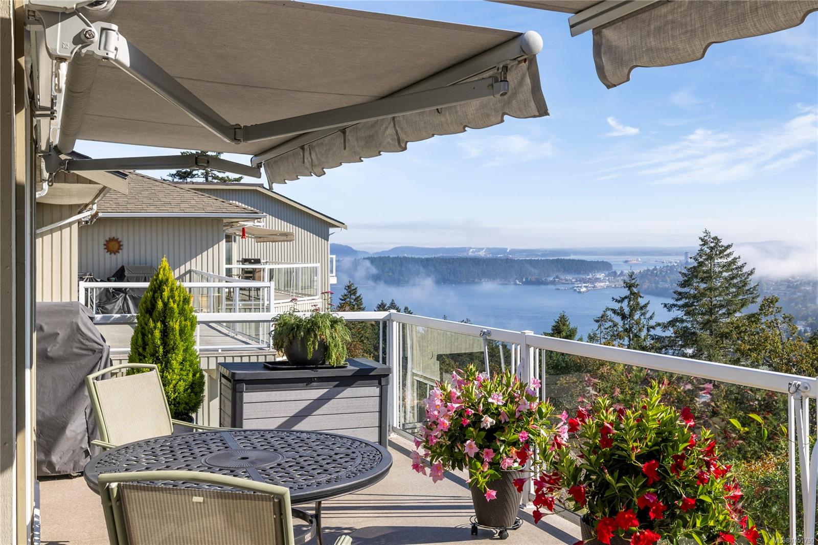 Furnished deck overlooking an inlet, with a retractable awning and colorful potted flowers.