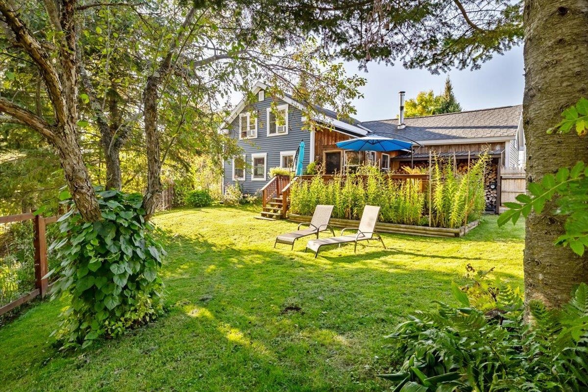 Blue-gray house behind a leafy backyard lawn, with a raised deck, garden beds, and lounge chairs.
