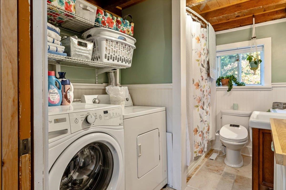Laundry area with washer and dryer beside a bathroom with a floral shower curtain and window plants.