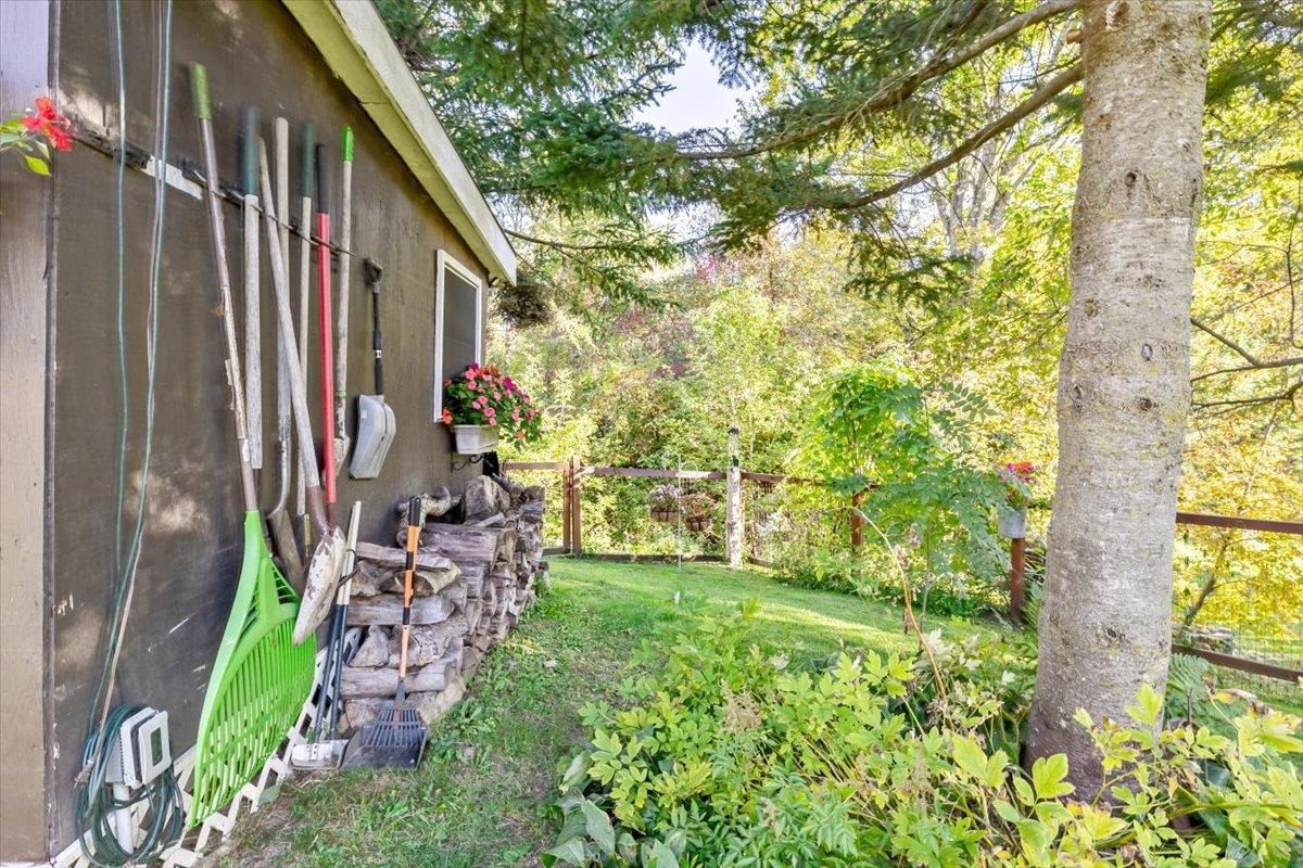 House side yard with garden tools, stacked firewood, a fenced lawn, and mature trees.