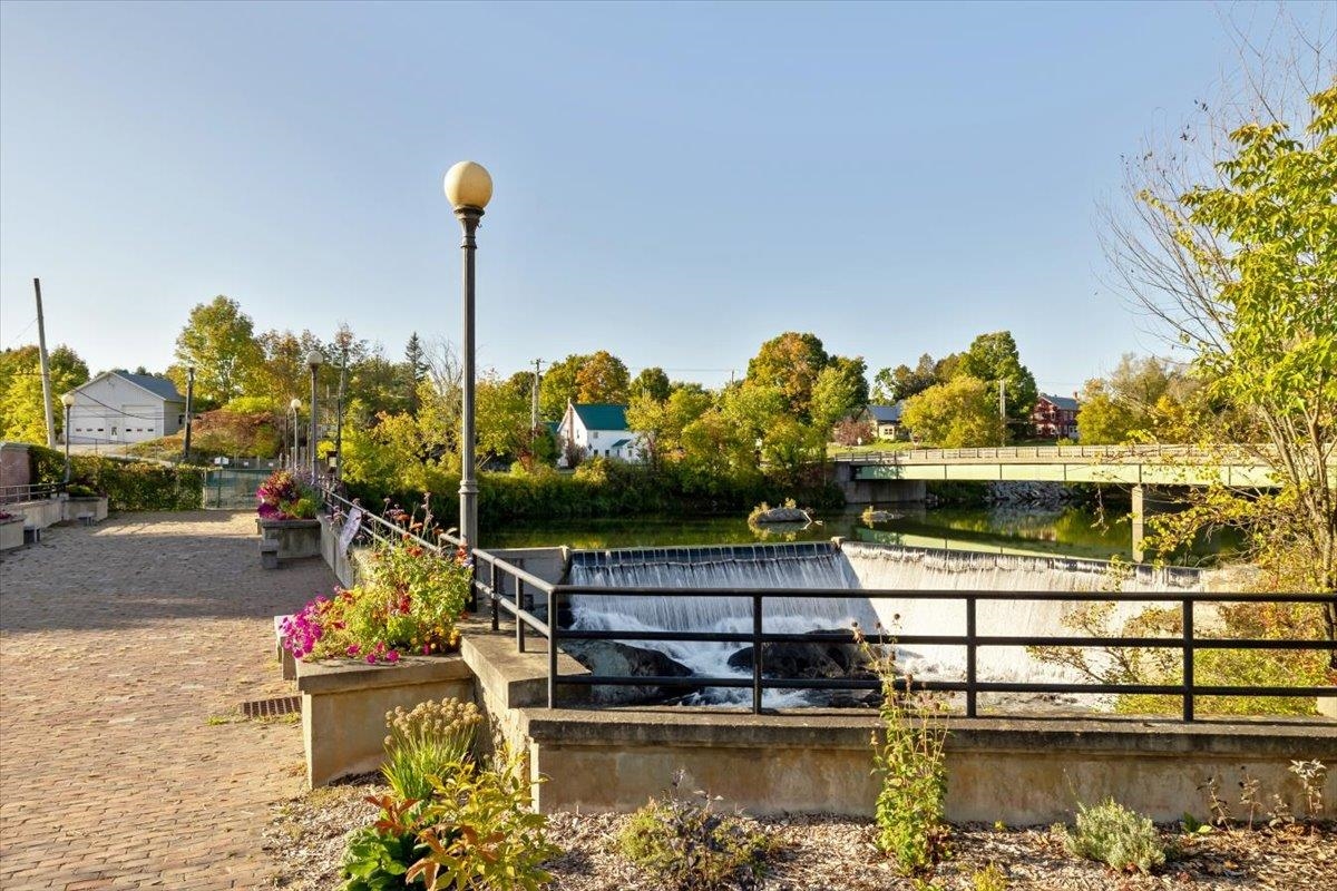 Riverside walkway beside a low waterfall, with flower planters and a bridge in the background.