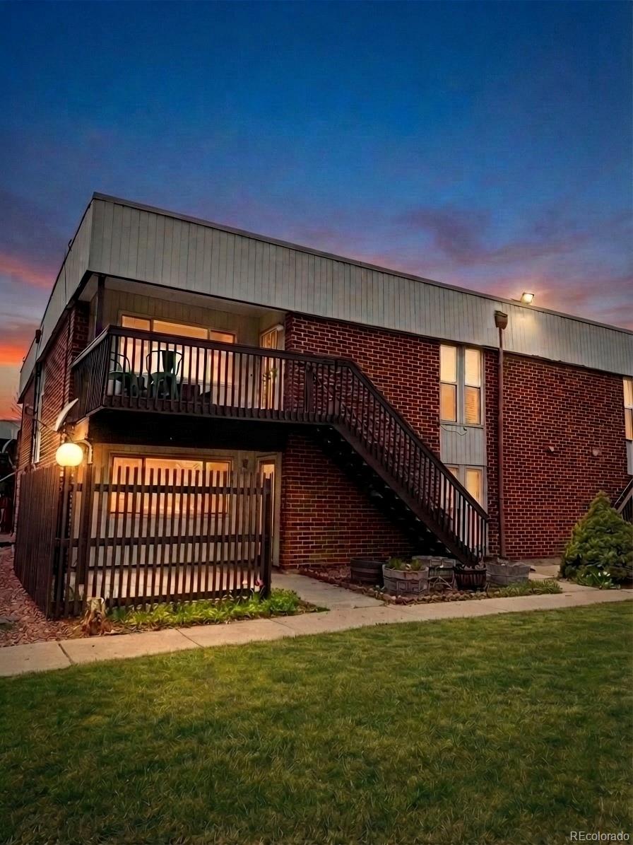 Brick residential building at dusk with an elevated balcony, exterior staircase, and lawn.