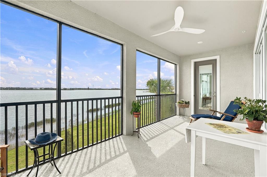 Covered balcony overlooking water, furnished with blue lounge chairs, potted plants, and a white table.
