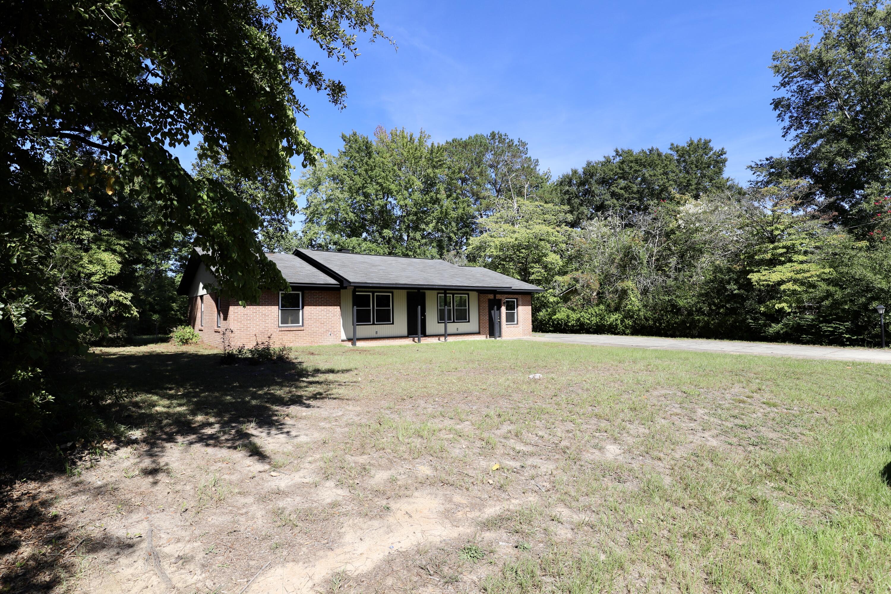 Single-story brick house with a covered porch, grassy yard, and surrounding mature trees.