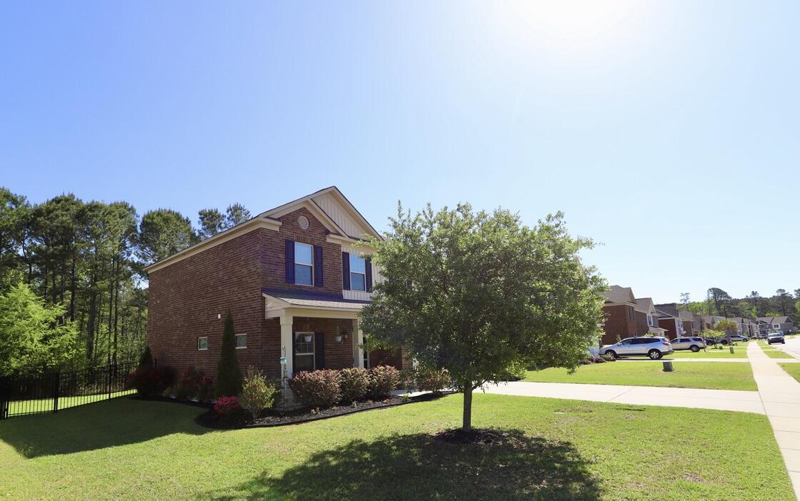 Two-story brick house with a covered porch, dark shutters, and a leafy tree in the front lawn.