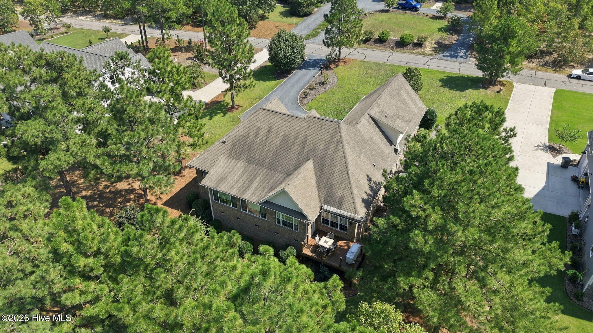 Aerial view of a brick home with a gray shingle roof, rear deck, and mature pine trees.