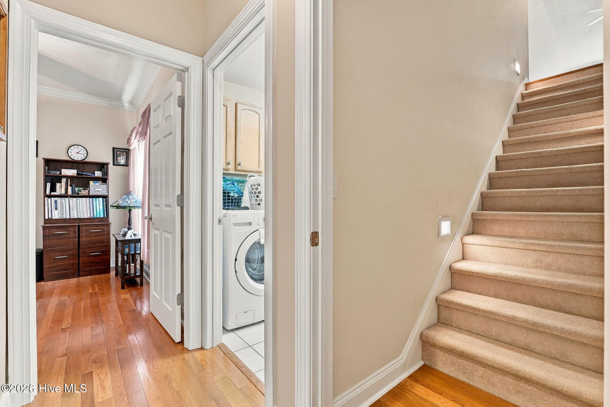 Hallway with hardwood floors, carpeted stairs, and open doorways to a book-lined room and laundry area.