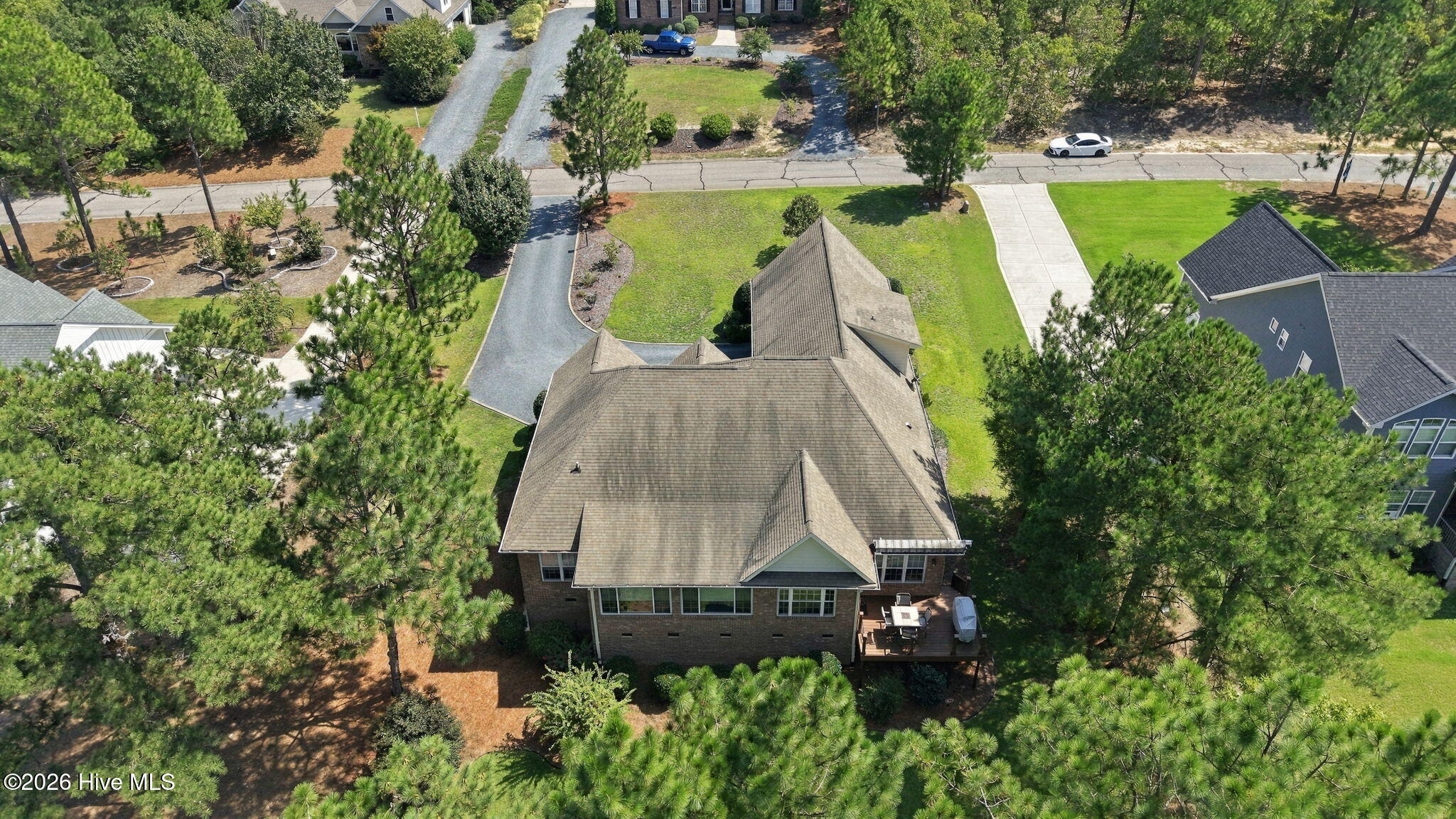 Aerial view of a brick home with a rear deck, broad roof, and pine-shaded landscaped yard.