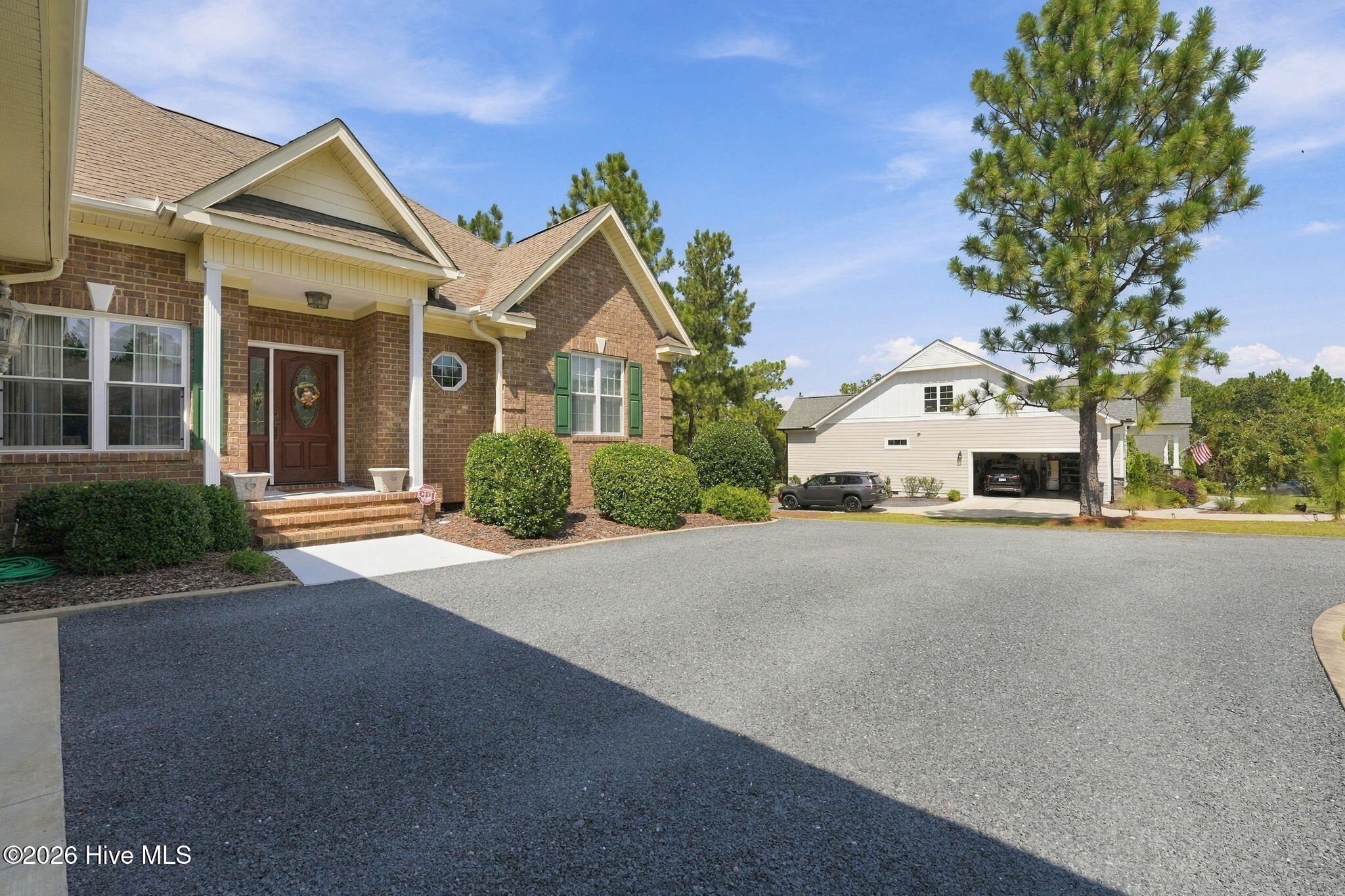 Brick home with covered entry and green shutters beside a broad driveway and tall pine tree.