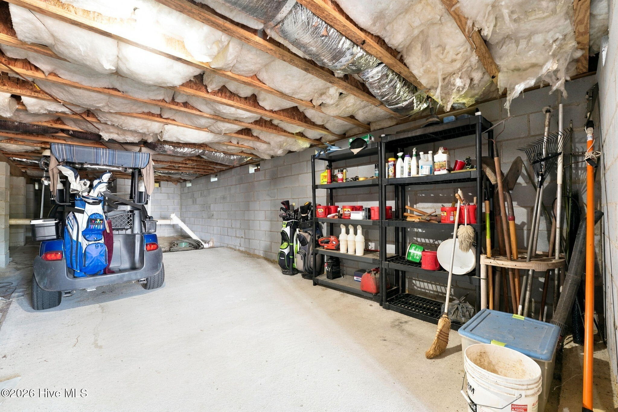 Garage with a golf cart, stocked shelving, golf bags, and gardening tools beneath exposed insulated joists.