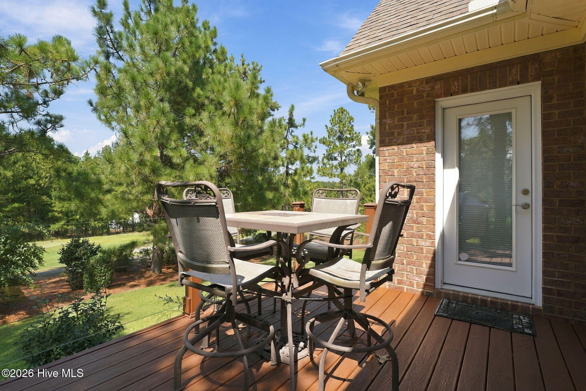 Furnished wooden deck with outdoor dining set overlooking a lawn and pine trees.