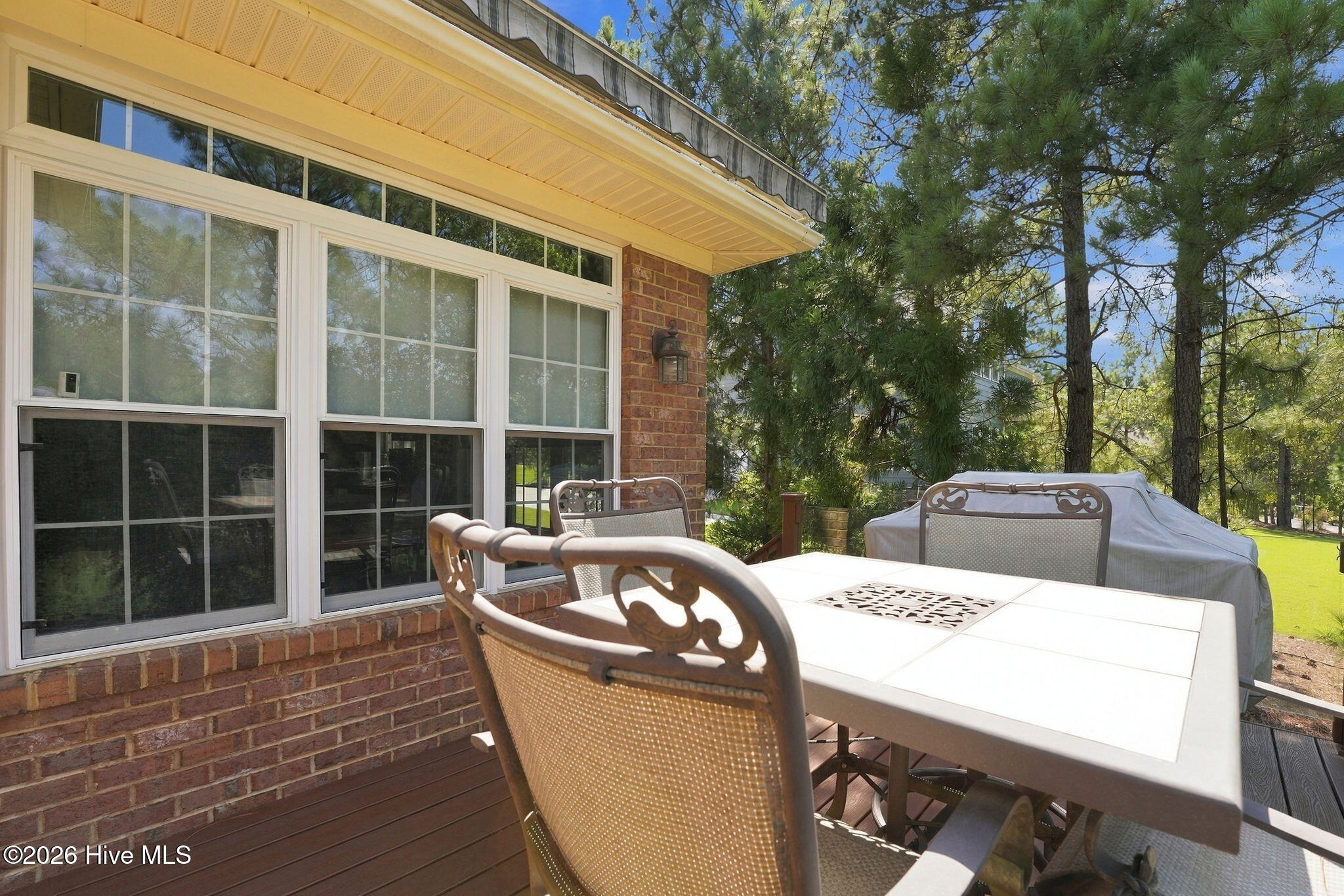 Wooden deck with outdoor dining set beside a brick house, surrounded by tall trees.